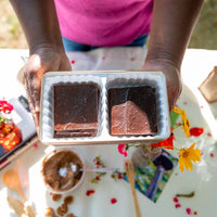Hands holding an open Koinonia Farm Dark Chocolate Espresso Bean Bark Gift Tin, showing two compartments of chocolate pecan bark above a table decorated with colorful flowers.