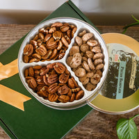 Open Koinonia Farm Pecan Sampler Gift Tin with three varieties of pecans in separate compartments, displayed on a green gift box with a gold ribbon, with the tin's lid beside it.
 Copy