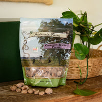 One-pound bag of Koinonia Farm Cinnamon Spiced Pecans on a rustic wooden table, with scattered pecans, a wicker basket, and green plants in the background.