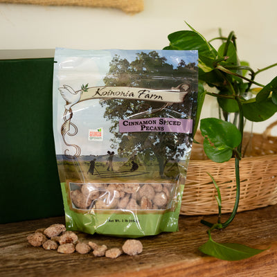 One-pound bag of Koinonia Farm Cinnamon Spiced Pecans on a rustic wooden table, with scattered pecans, a wicker basket, and green plants in the background.