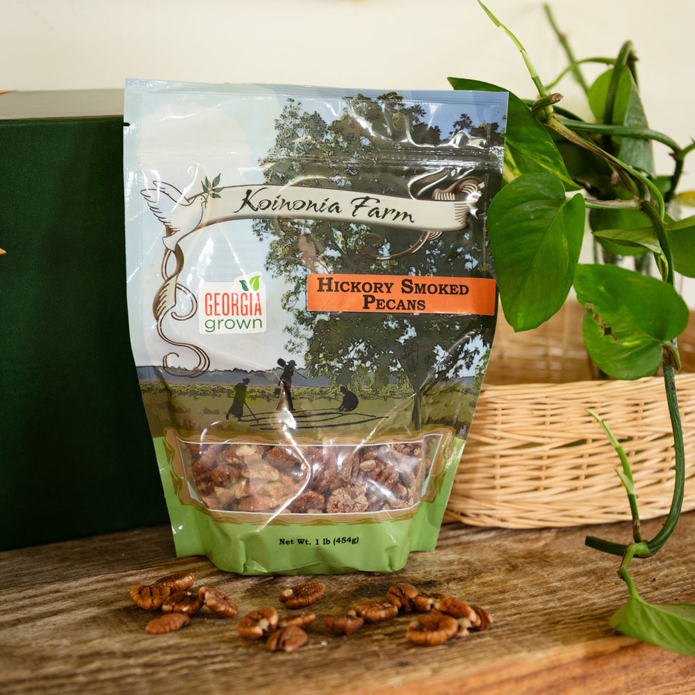 One-pound bag of Koinonia Farm Hickory Smoked Pecans on a rustic wooden table, with scattered pecans, a wicker basket, and green plants in the background.