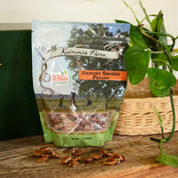 One-pound bag of Koinonia Farm Hickory Smoked Pecans on a rustic wooden table, with scattered pecans, a wicker basket, and green plants in the background.