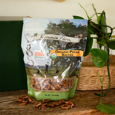 One pound bag of Koinonia Farm organic pecan halves displayed on a wooden table with greenery, a wicker basket, and scattered pecans.