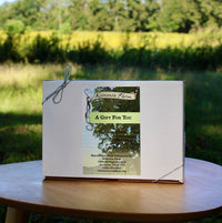 White Koinonia Farm displaying a label which says, A Gift for You. The gift box is tied with a thin silver ribbon, displayed outdoors on a wooden table, with a grassy field and trees in the background.